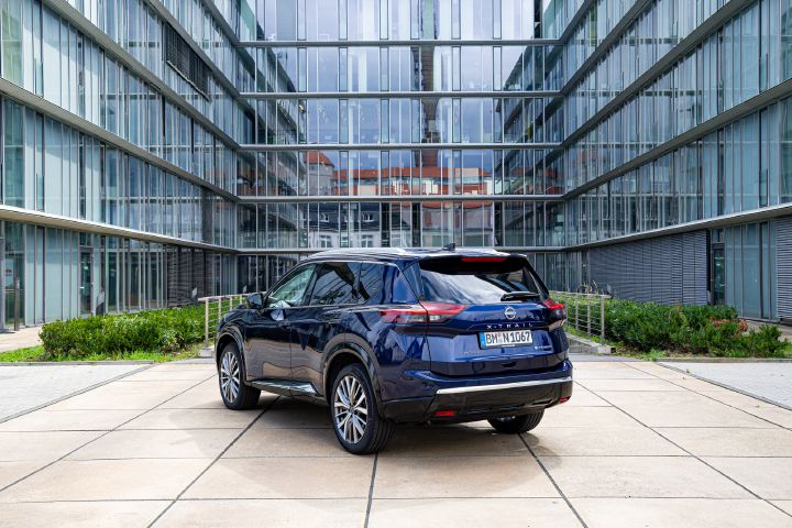 Ein dunkelblauer Nissan X‑Trail steht zwischen hohen Bürogebäuden mit gläsernen Fassaden, 3/4-Heckansicht