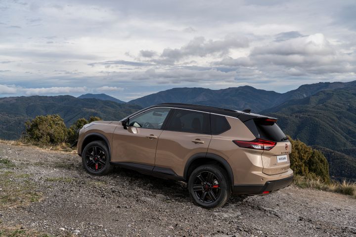 Der neue 2026 Nissan X‑Trail N-Trek in der Außenfarbe „Baja Storm” steht auf einem felsigen Hang mit Blick auf eine Bergkette unter bewölktem Himmel. Das Fahrzeug verfügt über schwarze Felgen mit magmaroten Bremssätteln und getönten Scheiben, die sein robustes Design und seine Geländegängigkeit unterstreichen.