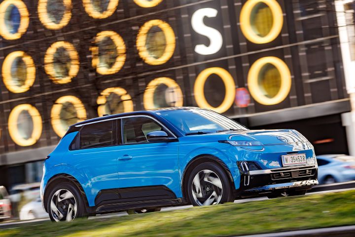 Ein leuchtend blauen Nissan Micra mit schwarzem Kontrastdach fährt seitlich an einem dunklen Gebäude mit hellen Lichtkreisen vorbei