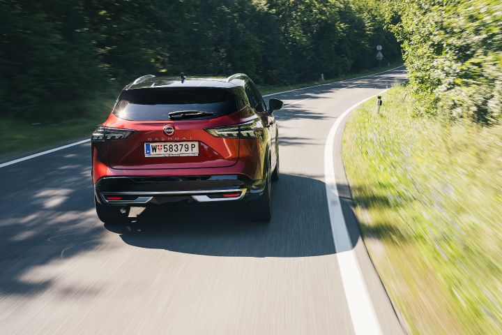 Ein Nissan Qashqai in Fuji Sunset Red fährt auf einer kurvigen Landtsraße mit dichtem Baumbestand an den Fahrbahnrändern, Heckansicht