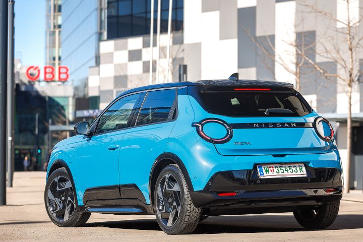 Statische 3/4-Heckansicht eines leuchtend blauen Nissan Micra mit schwarzem Kontrastdach vor einem Gebäude mit einer Fassade aus grauen und weißen Quadern und der Beschriftung ÖBB