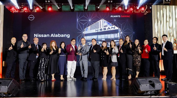 Group photo of the Nissan Alabang team with executives and guests on stage, celebrating their win at the 2025 Nissan Dealer of the Year Awards. Everyone is smiling and holding up the number one hand gesture. A large LED screen in the background displays 'Nissan Alabang' with an image of a dealership and the slogan 'Always Daring.