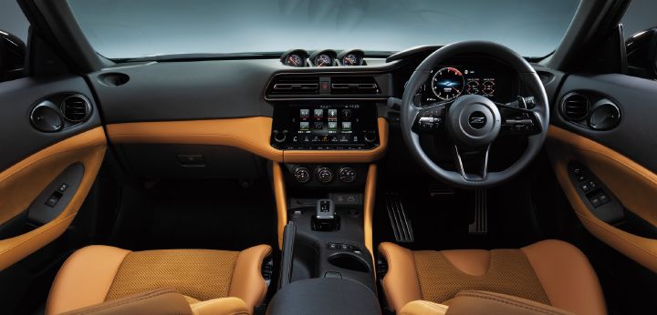 Cockpit view of a Fairlady Z featuring a tan interior. Photographed from the rear center of the cabin looking forward, the image shows a right-hand-drive layout with the steering wheel, digital instrument cluster, and central infotainment display positioned prominently. Tan leather upholstery is applied to the seats, dashboard trim, and door panels, creating a striking contrast with the black instrument panel. Three auxiliary gauges are mounted on top of the dashboard, emphasizing the sporty yet premium character of the interior.