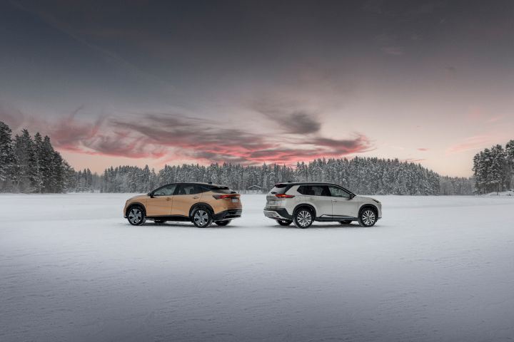 Elektroauto Nissan Ariya e‑4ORCE in Akatsuki Copper mit Dach und Außenspiegel in Pearl Black und silberner Nissan X‑Trail e‑Power e‑4ORCE stehen Heck an Heck auf schneebedecktem Eissee mit verschneitem Wald im Hintergrund, Seitenansicht