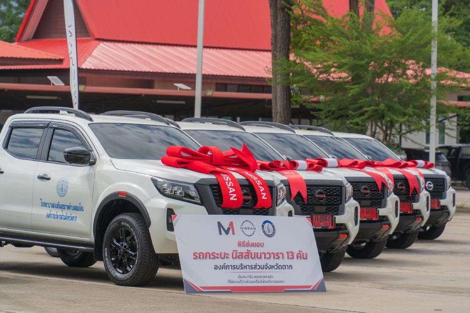 Several white Nissan pickup trucks decorated with red ribbons are lined up for an official vehicle handover ceremony.