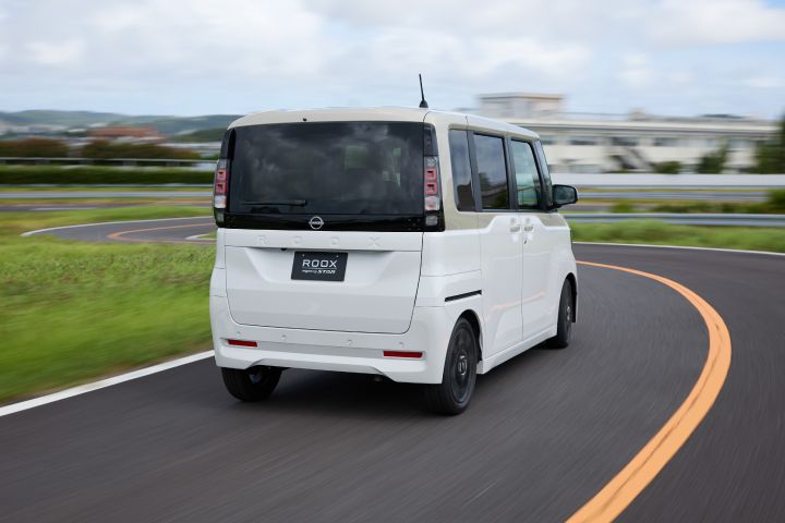 The new Nissan Roox kei car, in white with black wheels and a boxy design, is driving away on a winding road with green grass and a blue sky in the background.