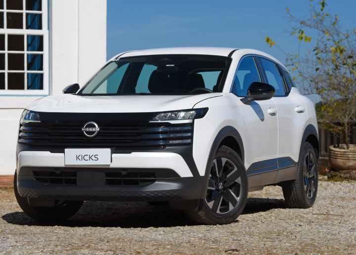 Novo Nissan Kicks Sense na cor branca em frente uma casa branca com janela, sobre o chão de brita e com o céu azul claro de fundo