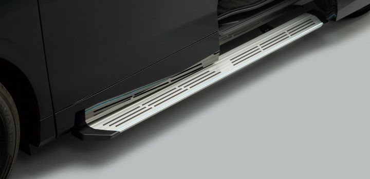 エルグランド ステップタイプの助手席側に装備された電動ロングステップを、車両側面やや前方からクローズアップで撮影した画像。ブラックのボディを持つ車両のスライドドアが開いた状態となっており、車体下部からシルバーの電動ロングステップが車外へ展開されている。ステップは車両に沿って横方向に長く伸び、表面には細長いスリット状の滑り止め加工が規則的に配置されている。
車体は光沢感のあるブラックで、左端にはタイヤの一部とサイドパネルが見える。開いたドアの奥にはブラックを基調とした車内空間がのぞき、フロアや乗降口周辺の内装が確認できる。ステップ外側の縁には淡い青白色のイルミネーションラインが点灯し、足元をやさしく照らしている。
