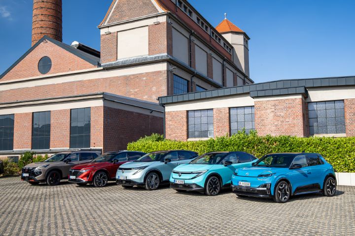 Vor einem Backsteingebäude und einer grünen Hecke stehen nebeneinander die fünf Nissan Modelle Nissan X‑Trail N-Trek, Nissan Qashqai, Nissan Ariya, Nissan Leaf und Nissan Micra, Blick von schräg vorne auf die Gruppe