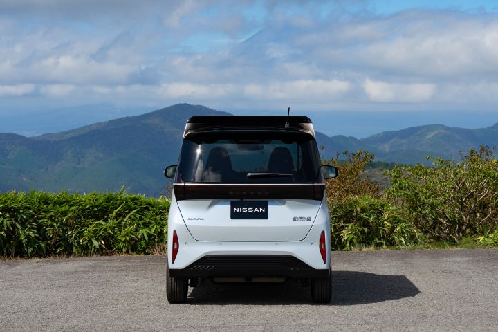 Under a blue sky, a light blue Nissan SAKURA is parked on the road, facing backward. The background includes green shrubs and distant mountains, with white clouds scattered across the sky.