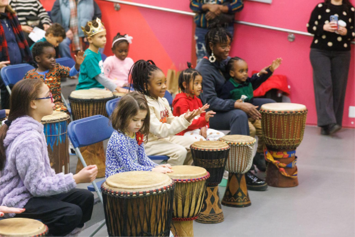A group of children site in a large group playing multiple different types of drum instruments.