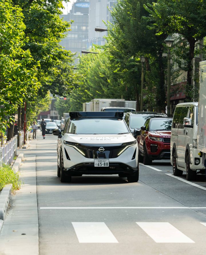 Ein weißer Nissan Ariya fährt auf einer städtischen Straße vorwärts, die von grünen Bäumen gesäumt ist, im Hintergrund sind mehrere Fahrzeuge und Gebäude zu sehen.