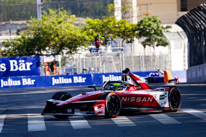 Oliver Rowland fährt mit dem Nissan Formel E Rennwagen Nr. 1 auf der Strecke in São Paulo und biegt nach rechts in eine Kurve ein, im Hintergrund grüne Blätter von den Bäumen und blaue Werbetafeln.