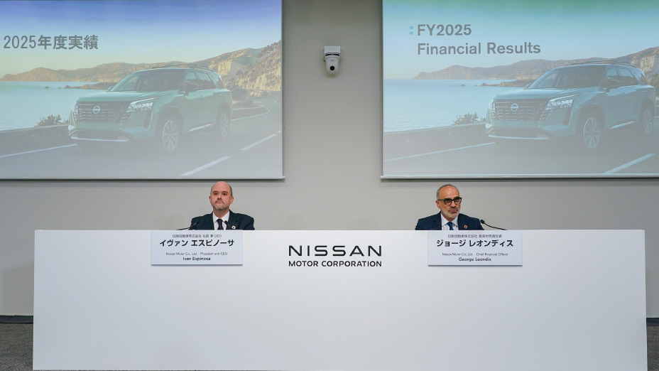 Eine Szene aus einer Pressekonferenz, in der zwei Personen nebeneinander vorne im Konferenzraum sitzen. Hinter ihnen sind auf Bildschirmen Bilder von Fahrzeugen und der Text „Geschäftsbericht 2025“ zu sehen, während an der Mittelwand das Logo der „NISSAN MOTOR CORPORATION“ zu erkennen ist