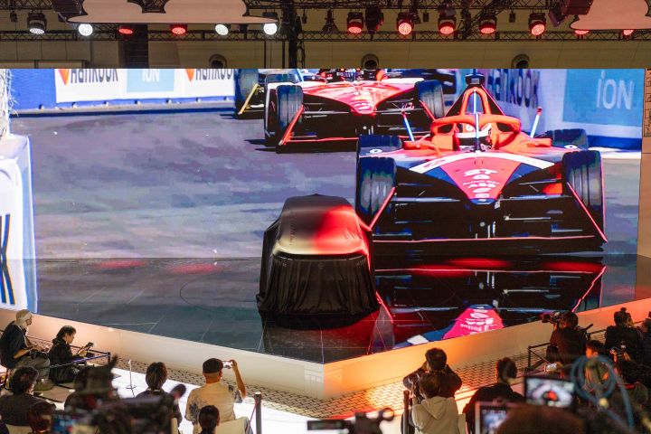 At the center of the stage, the all-new Elgrand is covered with a black cloth. In the background, a large screen displays an image of Formula E GEN3 cars racing. In the foreground, members of the press are gathered, taking photos with cameras.