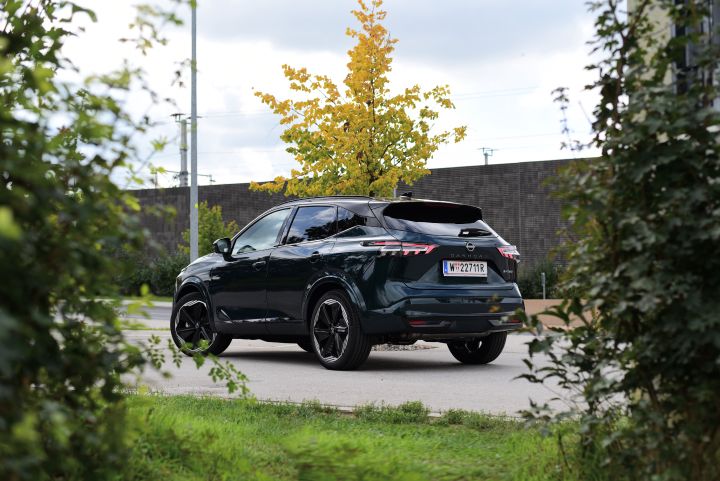 Zwischen zwei Büschen hindurch sieht man die 3/4 Heckansicht eines dunkelgrünen Nissan Qashqai e‑Power, der auf einem Parkplatz vor einer dunklen Mauer steht