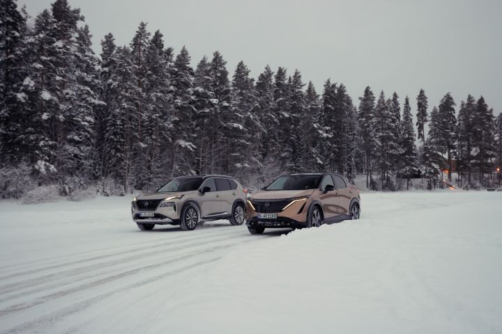 Silberner Nissan X‑Trail e‑Power e‑4ORCE und Elektroauto Nissan Ariya e‑4ORCE in Akatsuki Copper mit Dach und Außenspiegel in Pearl Black fahren nebeneinander auf schneebedeckter Straße mit verschneitem Wald im Hintergrund, 3/4 Front