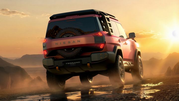 A red Terrano PHEV Concept seen from a rear three-quarter view, driving to the right across a desert landscape at sunset. Sandy and rocky terrain surrounds the vehicle, with reflections on wet ground and distant mountains under the setting sun.