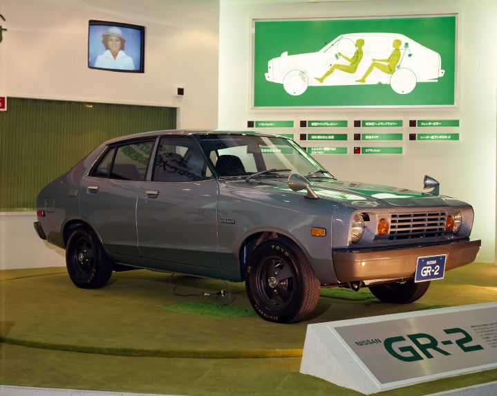 a light gray GR-2 vehicle on display at the auto show. in the background there is a green graphic showing 2 mannequin figures seated inside the vehicle.