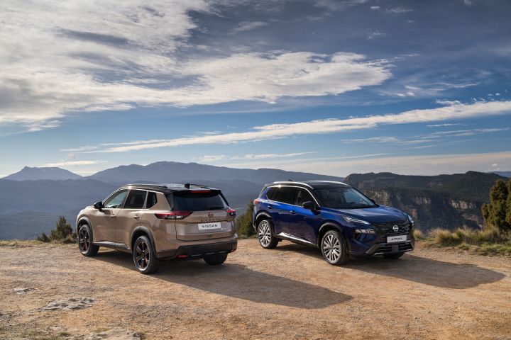 Deux nouveaux Nissan X‑Trail, le  Tekna Bleu Sukumo et le N-Trek Sable Miho, sont stationnés sur une clairière de terre, avec un paysage montagneux et un ciel partiellement nuageux en arrière-plan.