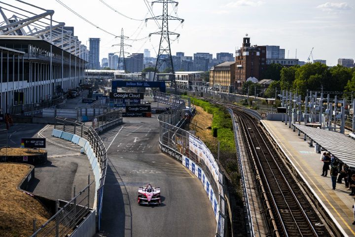 Ein rot-weißer Nissan Formel-E-Rennwagen fährt auf einem eingezäunten Straßenkurs in London vorwärts. Rechts neben der Strecke verlaufen Bahngleise und ein Bahnsteig, im städtischen Hintergrund sind Gebäude und Stromleitungen zu sehen.