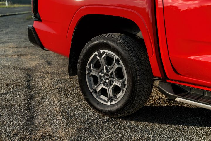 Close-up of the rear wheel and lower rear quarter of a 2026 Nissan Navara ST-X in Outback Red, showing the alloy wheel, tyre tread, mud flap, rear bumper corner, and side step.