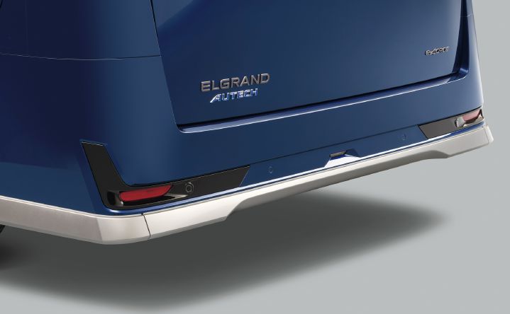 ブルーのエルグランド AUTECHのリヤ下部を、左後方からクローズアップで撮影した画像。車両後端のバックドア左下にはシルバーの「ELGRAND」エンブレムと、ブルーの「AUTECH」エンブレムが配置されており、右下には「e‑4ORCE」バッジが見える。車両は右方向を向いており、リア左側から右側にかけてのデザインが確認できる構図となっている。
リヤバンパー下部には、AUTECH専用デザインのメタル調フィニッシュのプロテクタが装着され、ボディカラーのブルーとのコントラストを形成している。左右下端には赤色のリフレクターが配置され、その周囲にはブラックのアクセントパネルと円形のセンサーが組み込まれている。バックドアおよびバンパー表面には光沢があり、滑らかで上質な質感が強調されている。