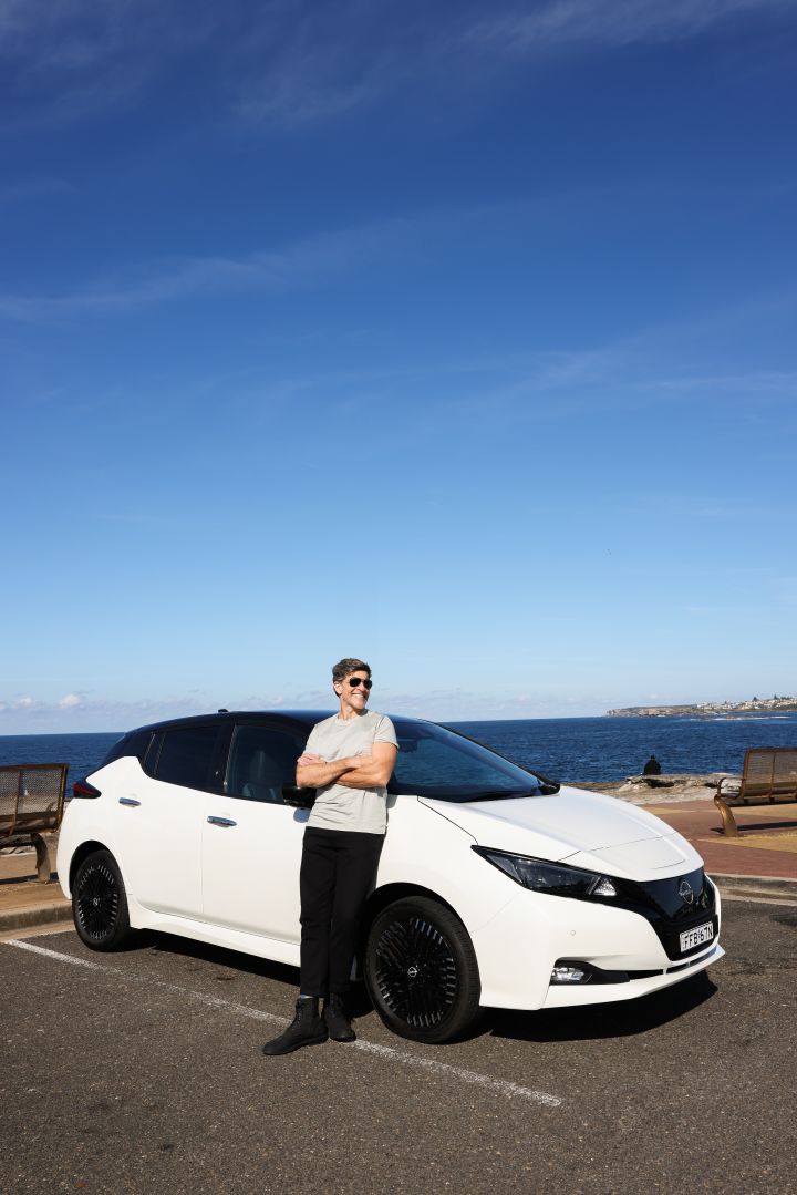 Nissan Australia ambassador Osher Gunsberg leans on his white Nissan Leaf parked in front of a beach