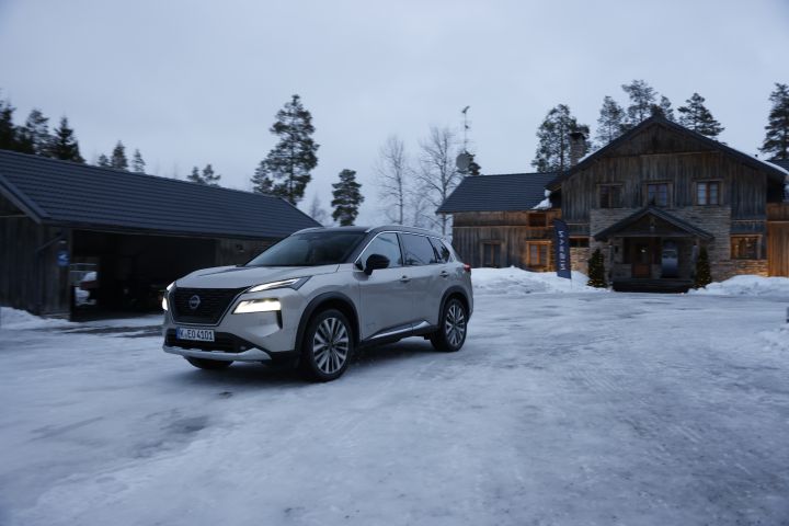 In der Abenddämmerung fährt ein silberner Nissan X‑Trail e‑Power e‑4ORCE auf Schnee vor einem großen Holzhaus