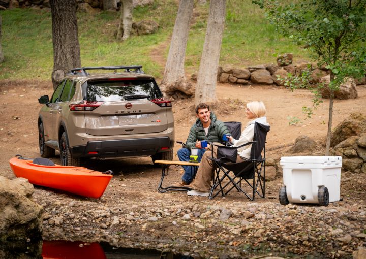 The 2025 Nissan rogue rock creek backed in near a creek with two people sitting with a cooler, and red Kayak.