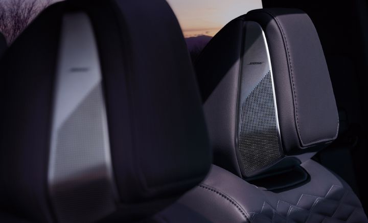 Close-up interior image of the All-new ELGRAND headrests, highlighting the integrated BOSE headrest speaker system. The black leather-style seat headrests feature metallic-finish speaker panels with the BOSE logo, built into the sides of the headrests. Two headrests are visible, with the rear headrest speaker in sharp focus and the foreground headrest softly blurred. Decorative stitching and geometric quilted patterns on the seat upholstery enhance the premium appearance. Through the window, a sunset sky and silhouettes of trees can be seen in the background, while the subdued lighting emphasizes the vehicle’s upscale audio and interior craftsmanship.