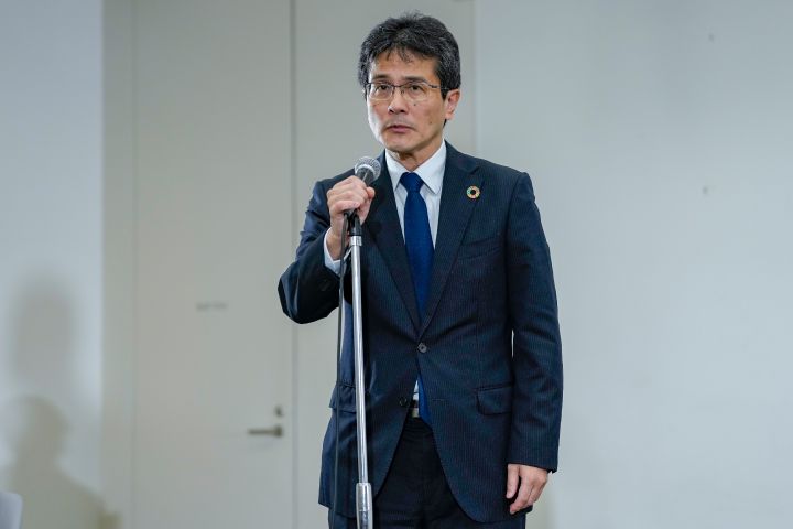 スーツを着た日産自動車 執行役、チーフ モノづくり オフィサーである ひらた ていじ がマイクの前で話している。記者会見の様子を示している。