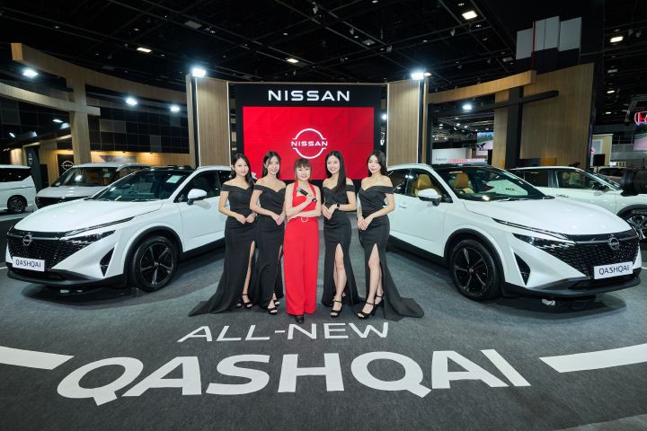 A lady in red dress with Nissan ladies stand between the two white-color Nissan Qashqai displayed. The red Nissan logo on LED screen at the back