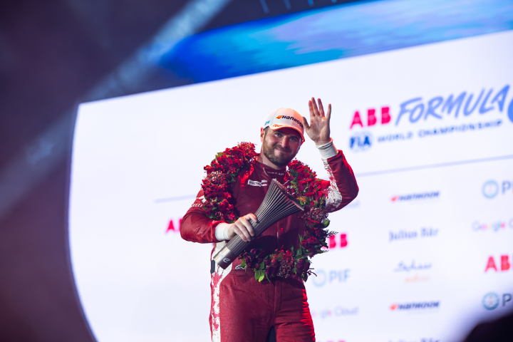 Oliver Rowland vom Nissan Formula E Team trägt seinen roten Rennanzug, hält in seiner rechten Hand die Trophäe für den dritten Platz beim E-Prix in Dschidda und winkt mit der linken Hand der Menge zu, auf dem Kopf trägt er eine weiße Kappe.