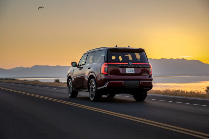 Left rear 2/4 angle of a 2025 Nissan Armada driving on a road during sunset with a mountain and body of water behind.