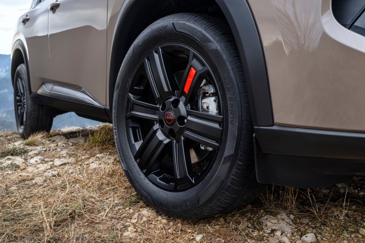Der neue 2026 Nissan X‑Trail N-Trek in der Karosseriefarbe „Baja Storm“. Nahaufnahme eines schwarzen Leichtmetallrads und Reifens, geparkt auf felsigem Gelände. Das Rad verfügt über einen magmaroten Bremssattel und ein zentrales Logo, die die Geländegängigkeit und die robusten Designelemente unterstreichen.