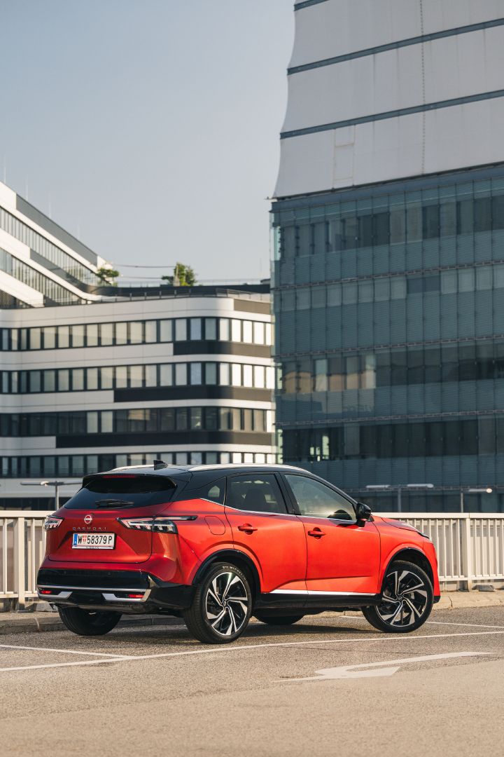 3/4 Heckansicht eines Nissan Qashqai in Fuji Sunset Red auf einem Parkplatz vor Hochhäusern