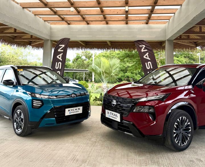 Front view of a blue Nissan Kicks e‑POWER and a red Nissan X‑Trail e‑POWER displayed side by side under a covered outdoor pavilion, with Nissan-branded flags and lush greenery in the background. Visible vehicle badges identify the models as Kicks e‑POWER and X‑Trail e‑POWER