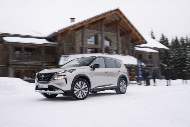Silberner Nissan X‑Trail e‑Power e‑4ORCE fährt auf schneebedecktem Untergrund mit Menschen und großem Holzhaus im Hintergrund