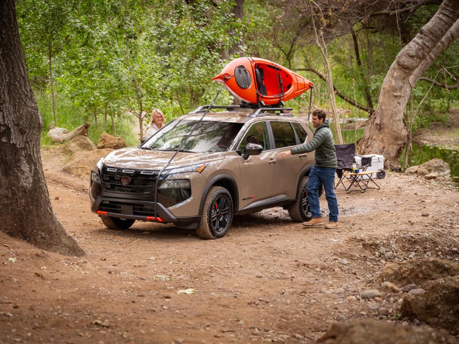 Front left 3/4 angle of a 2025 Nissan Rogue parked on a camp site with a kayak on its roof and a man opening the door.