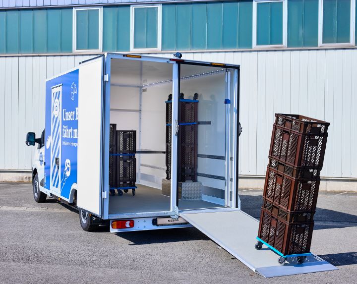 Blick von schräg hinten auf den Nissan Interstar-e mit Bäckereikoffer, auf der heruntergeklappten Laderampe steht ein Wagen mit braunen Bäckereikörben, um ins Innere geschoben zu werden