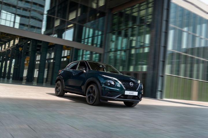 The Nissan JUKE Pulse driving through a modern urban setting, shown in motion with a sleek Ocean Deep exterior against glass-fronted buildings.