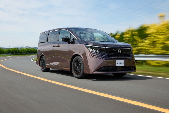 新型エルグランドが、左から右へ向かってカーブした舗装道路を走行している。車両は右前方へ進む斜め前からのアングルで捉えられており、ブラックのグリルと精細なLEDヘッドライトが強調されている。背景には青空と薄い雲が広がり、左右には緑の植生がぼかし表現で流れるように描かれ、走行中のスピード感が表現されている。