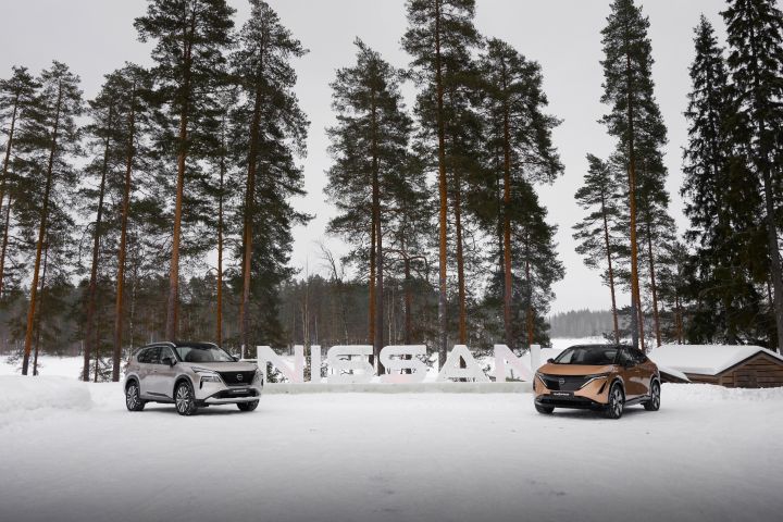Silberner Nissan X‑Trail e‑Power e‑4ORCE und Elektroauto Nissan Ariya e‑4ORCE in Akatsuki Copper mit Dach und Außenspiegel in Pearl Black stehen auf schneebedecktem Untergrund, zwischen ihnen steht ein weißer Nissan-Schriftzug