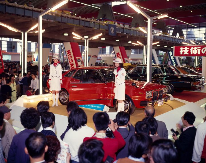 a large crowd of people surrounding a display of a red vintage car at the auto show, with 2 female presenters dressed in white and red speaking on a microphone
