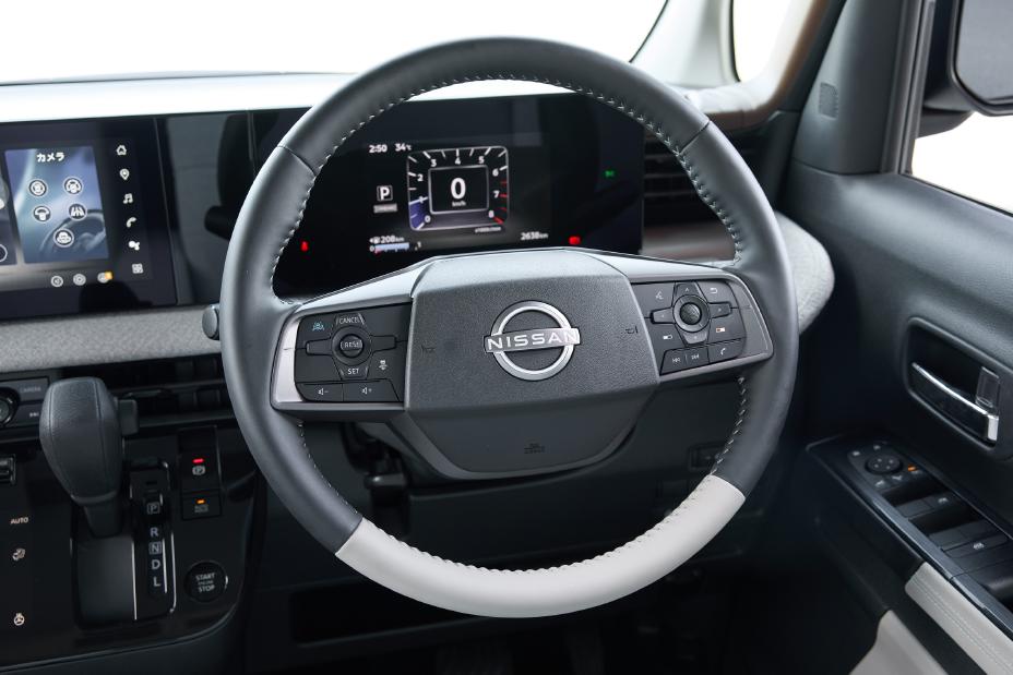 The driver's seat of the new Nissan Roox kei car. The steering wheel features the Nissan logo at the center, surrounded by various control buttons. Digital displays and an infotainment system are visible in the background.