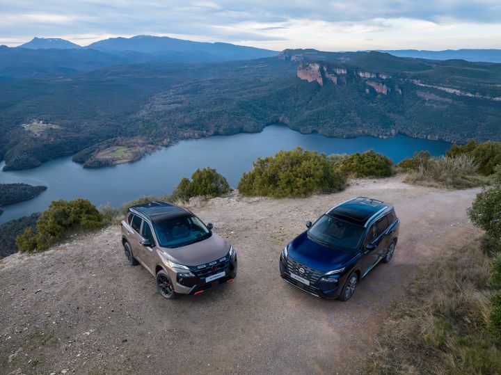 Zwei neue Nissan X‑Trail des Modelljahres 2026 stehen auf einem unbefestigten Aussichtspunkt mit einem großen See und bewaldeten Bergen im Hintergrund. Die Fahrzeuge, ein N-Trek in der Karosseriefarbe Baja Storm und ein Tekna in der Karosseriefarbe Universal Blue, stehen nebeneinander auf einem unebenen Gelände mit Büschen und klarem Himmel darüber.