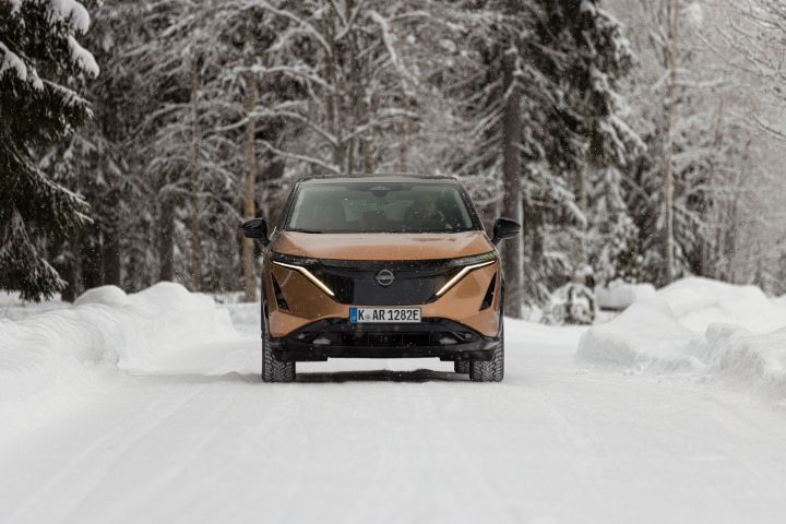 Elektroauto Nissan Ariya e‑4ORCE in Akatsuki Copper mit Dach und Außenspiegel in Pearl Black fährt auf schneebedeckter Straße mit verschneiten Bäumen auf beiden Seiten, Front
