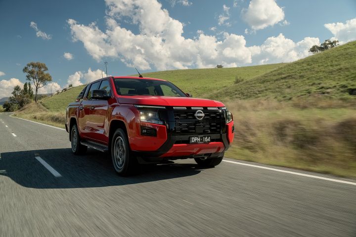 Front three-quarter view of a 2026 Nissan Navara ST‑X Outback Red dual cab driving along a sealed rural road surrounded by farmland and distant rolling hills.