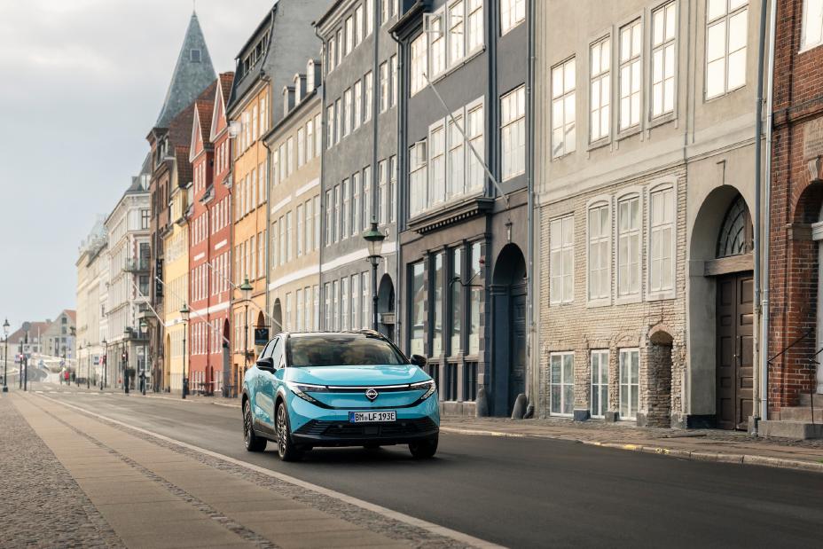 Ein türkiser Nissan LEAF fährt leicht schräg auf einer städtischen Straße in Kopenhagen vor bunten Stadthäusern